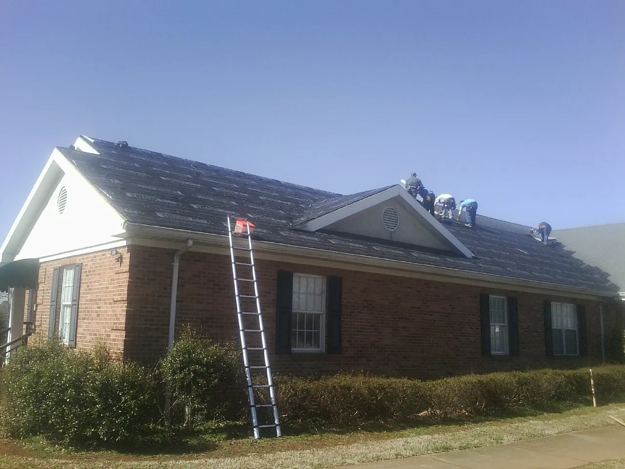 Crew tearing shingles off a brick ranch roof with a ladder set — Emergency Roof Repair work by our crew in Bardolph